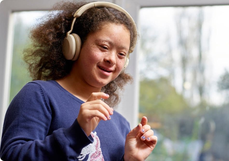 A smiling girl with curly hair wears headphones and dances with her hands raised in front of a window.